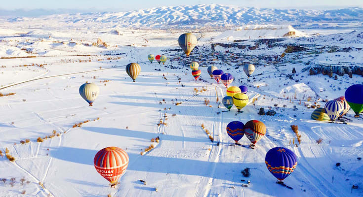 Multiple colorful hot air balloons floating over snowy winter landscape in Cappadocia, Turkey with mountains in background
